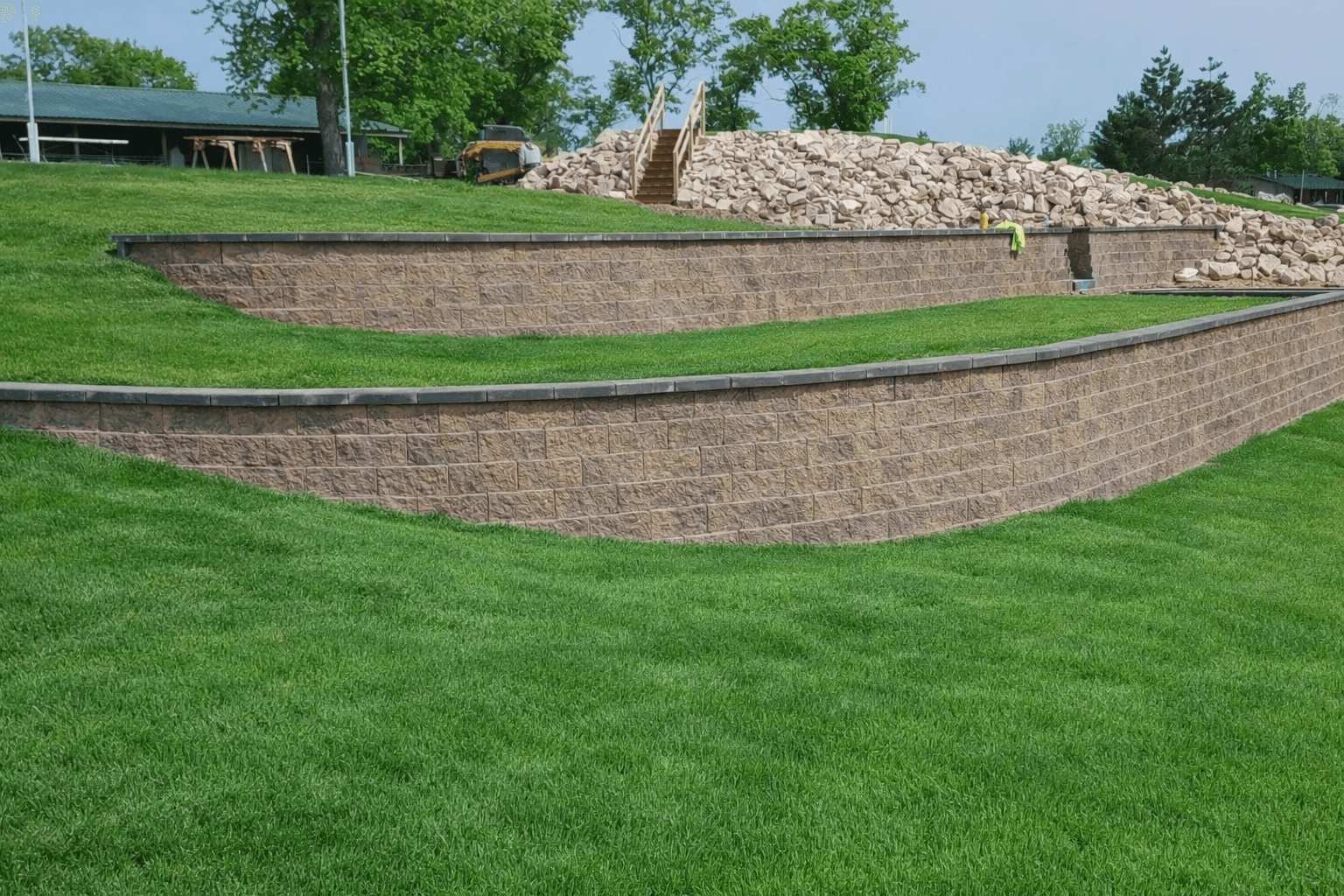 Multi-tiered curved segmental block retaining walls in tan with dark gray cap stones, professional sod installation on terraced levels, commercial landscaping with erosion control and elevation management, yellow excavator staging limestone boulders, commercial building with green roof, Wisconsin commercial landscape construction